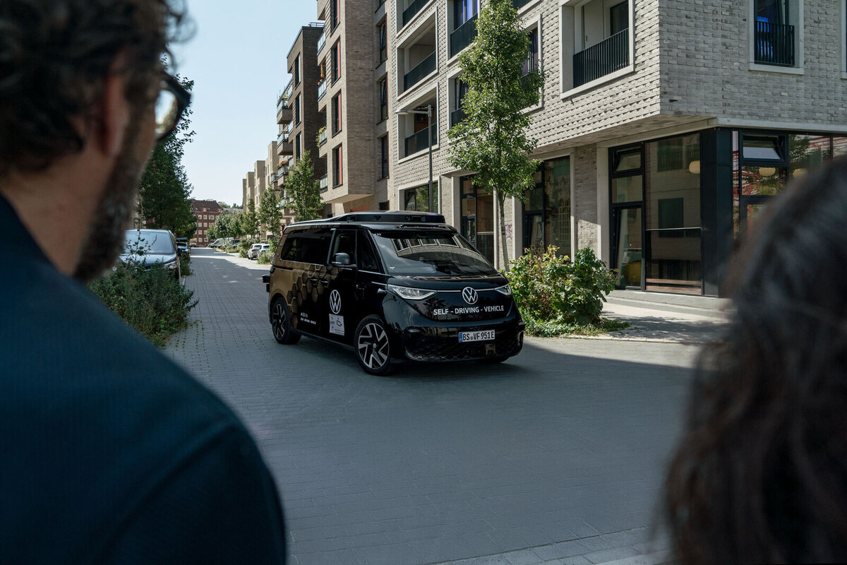Self-driving VW vehicle driving on an urban road while cyclists and pedestrians move along the side.