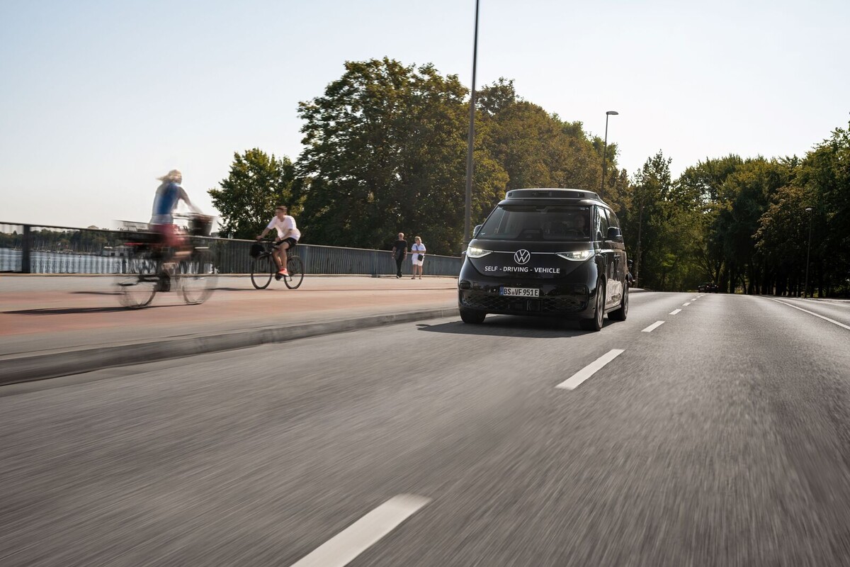 Self-driving VW vehicle driving on an urban road while cyclists and pedestrians move along the side.